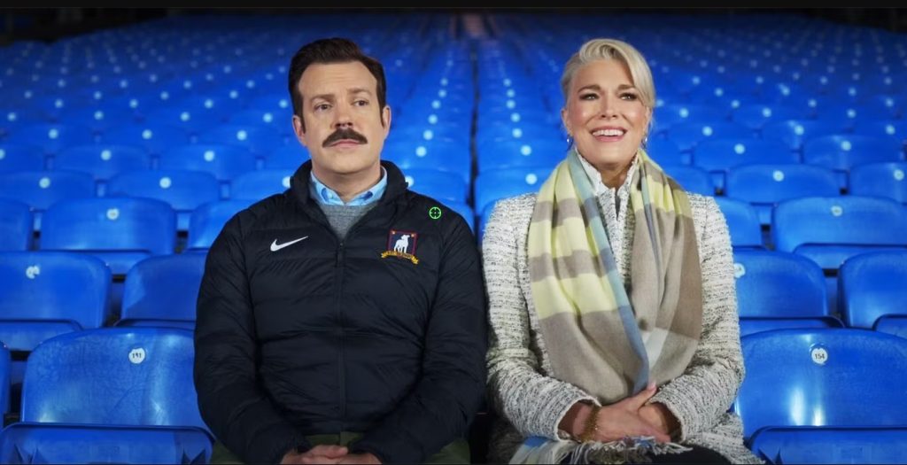 Ted Lasso and Rebecca Welton sit side by side in the empty stands of AFC Richmond's stadium, sharing a quiet moment in Ted Lasso.