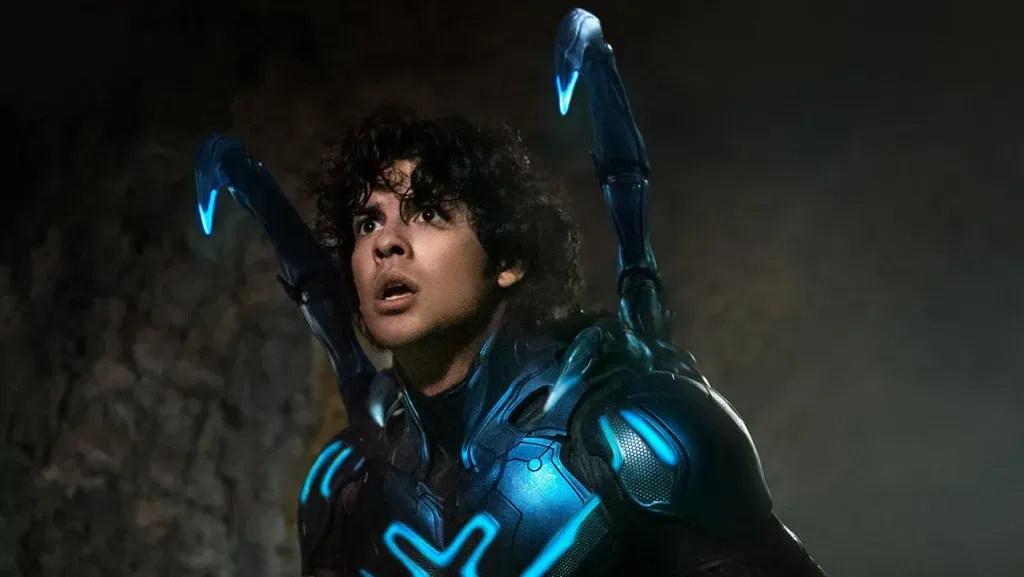 A young superhero in a glowing blue armored suit with mechanical beetle-like appendages looks shocked inside a dimly lit cave. Blue Beetle earned more than Supergirl