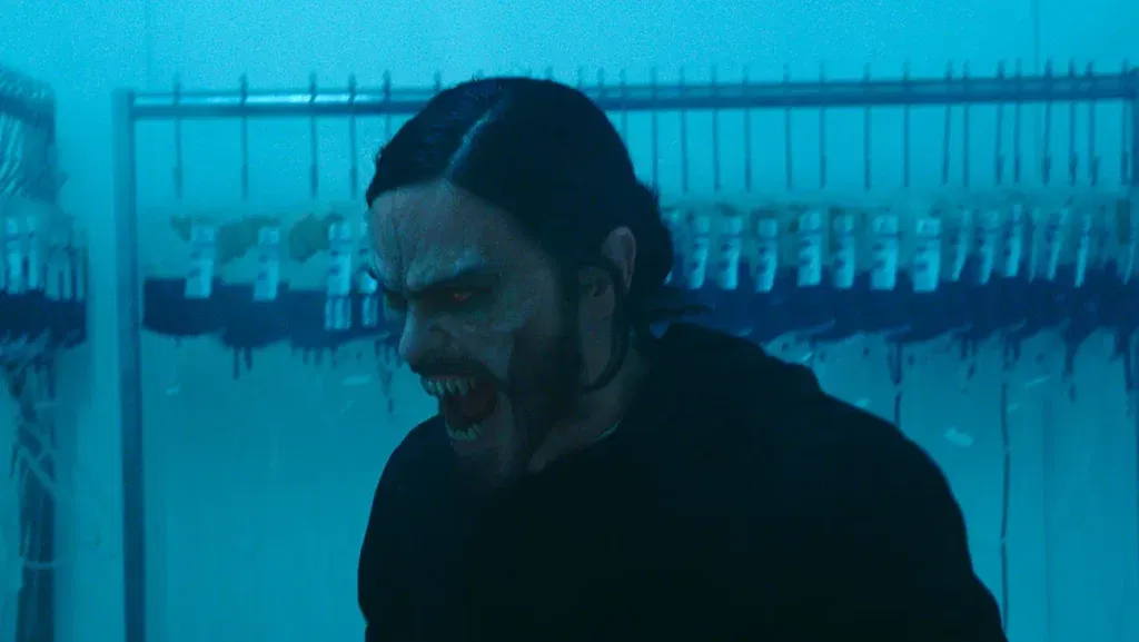 A pale, monstrous figure with glowing red eyes and sharp fangs snarls inside a dimly lit laboratory in a scene from Morbius.