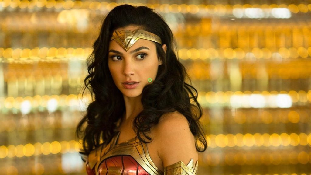 Wonder Woman, wearing her iconic tiara and armor, looks over her shoulder in a brightly lit indoor setting in Wonder Woman 1984.