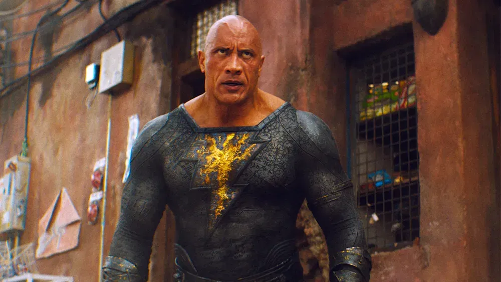 Black Adam stands in his black suit with a glowing lightning emblem, staring ahead in a scene from Black Adam (2022).
