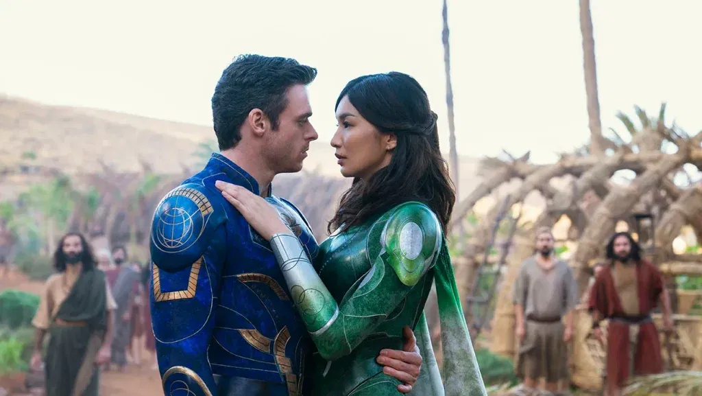 Two Eternals stand face to face in their blue and green armor while embracing in a village setting in Eternals.