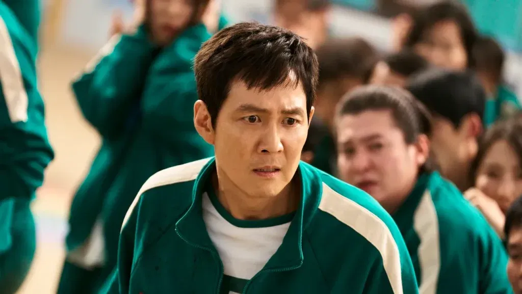 A man in a green tracksuit looks distressed as he stands among other contestants in matching uniforms in Squid Game.