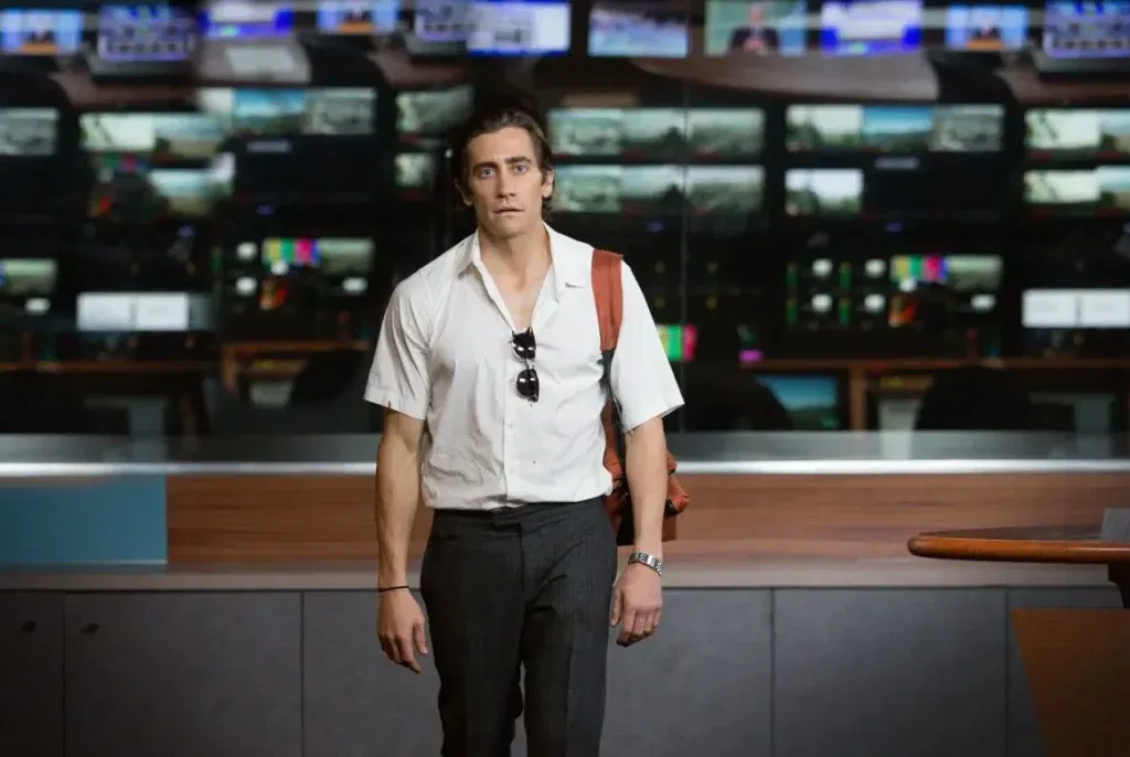 A man stands inside a television newsroom surrounded by monitors, wearing a white shirt and carrying a brown shoulder bag. This is a still from the movie Nightcrawler and features Jake Gyllenhaal