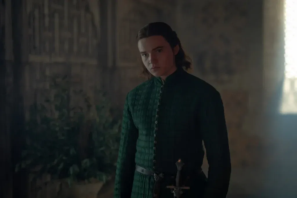Daeron Targaryen standing inside a dimly lit castle room in House of the Dragon, dressed in dark green Targaryen clothing with a sword at his side.