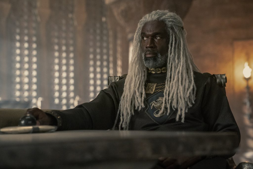 Corlys Velaryon sits at a table inside a dimly lit castle chamber, wearing dark armor and a gold necklace, with his long silver-gray dreadlocks framing his face.