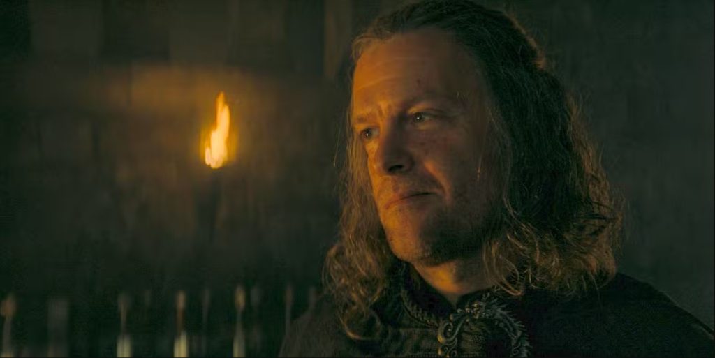 Ulf White sits in a dimly lit room, illuminated by torchlight, wearing dark clothing and looking off to the side.