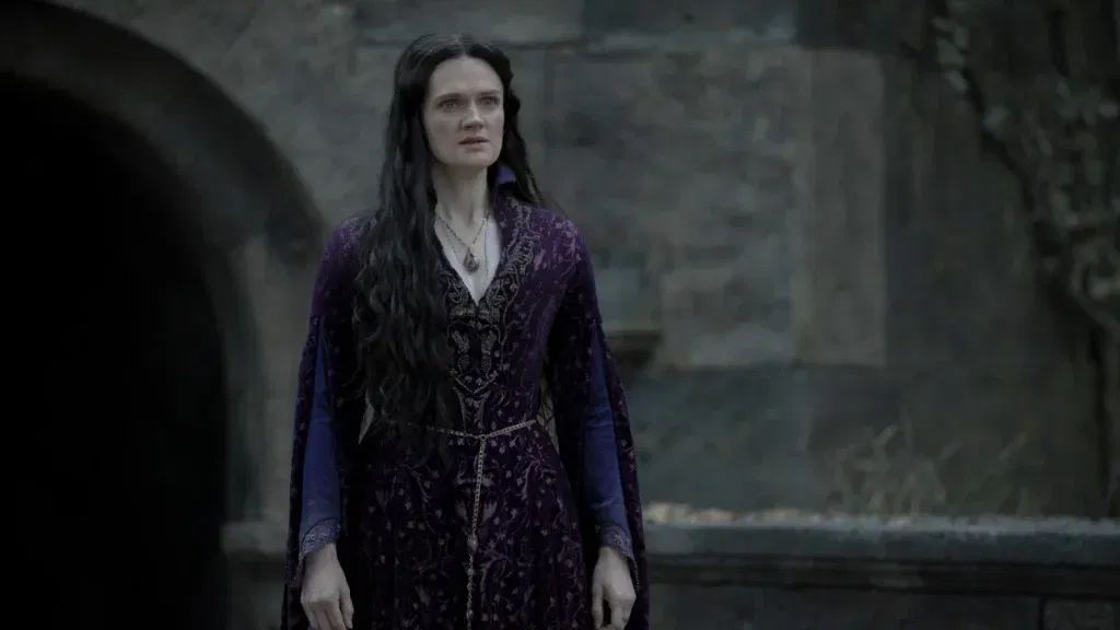 Alys Rivers stands in a stone courtyard wearing a dark purple gown, with long black hair and a pendant around her neck.