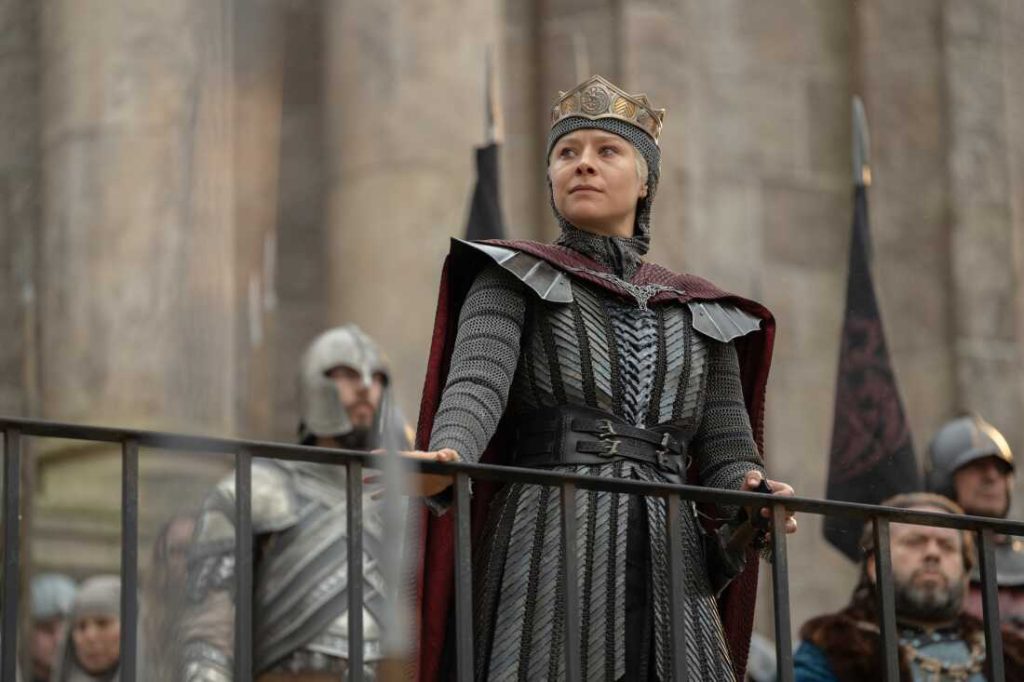 Rhaenyra Targaryen in House of the Dragon season 3 stands on a castle balcony wearing a crown and black armor with a red cloak, surrounded by soldiers.