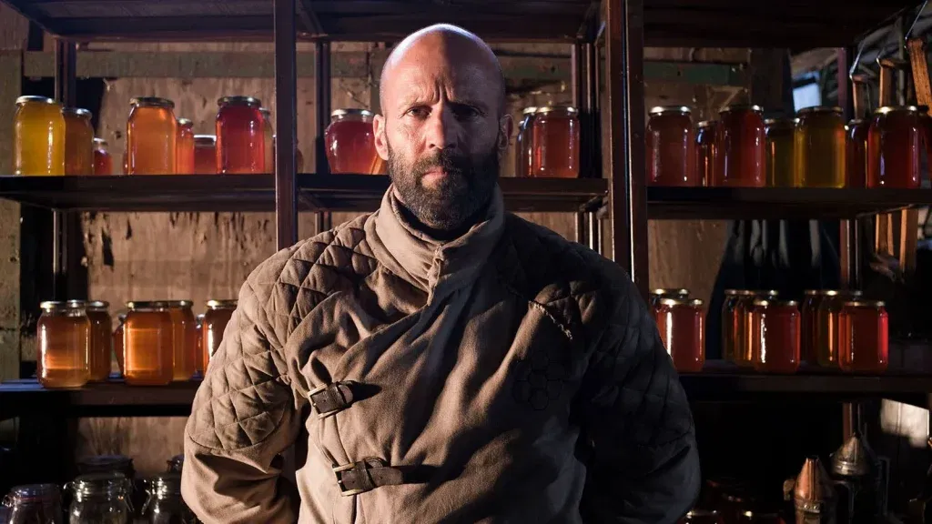 Jason Statham in a quilted beekeeping suit standing before shelves stacked with jars of honey.