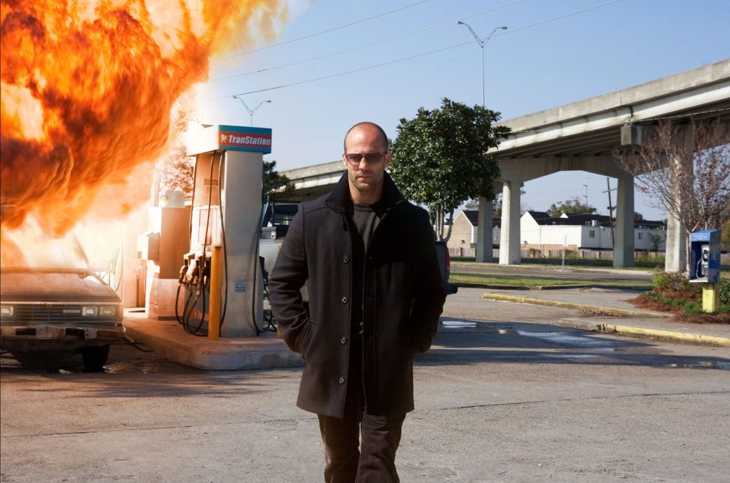 Jason Statham in a dark coat and sunglasses walking away from a gas station engulfed in flames, with a laser sight visible on his chest.