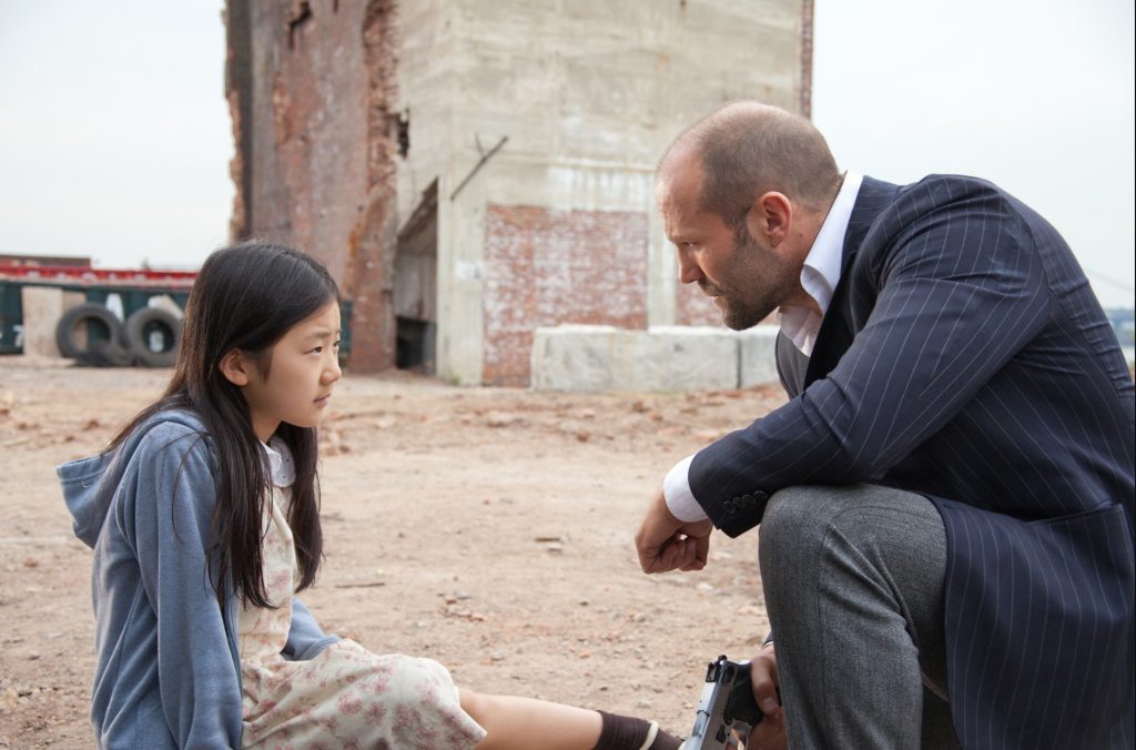 Jason Statham in a pinstripe suit crouching with a gun to speak with a young girl in a hoodie and floral dress on a demolition site.