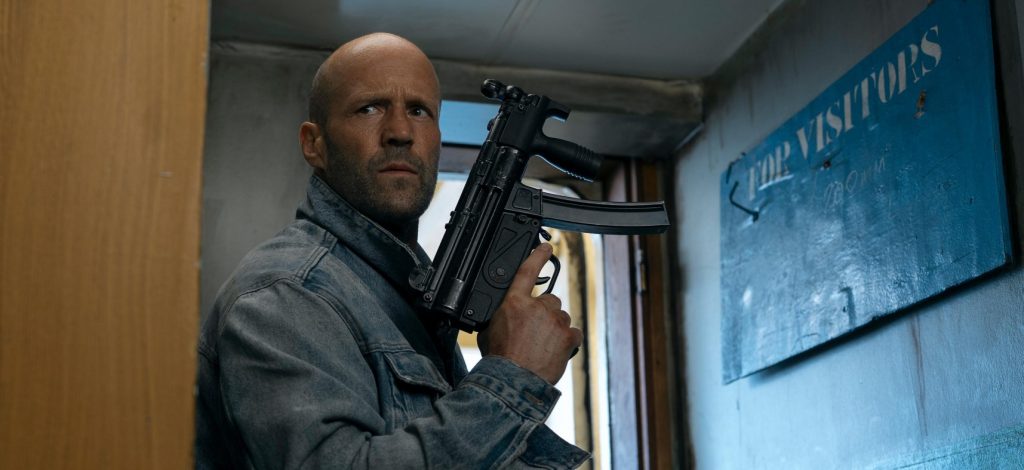 Jason Statham in a denim jacket holding a submachine gun while pressed against a wall in a narrow corridor, with a "For Visitors" sign visible beside him.