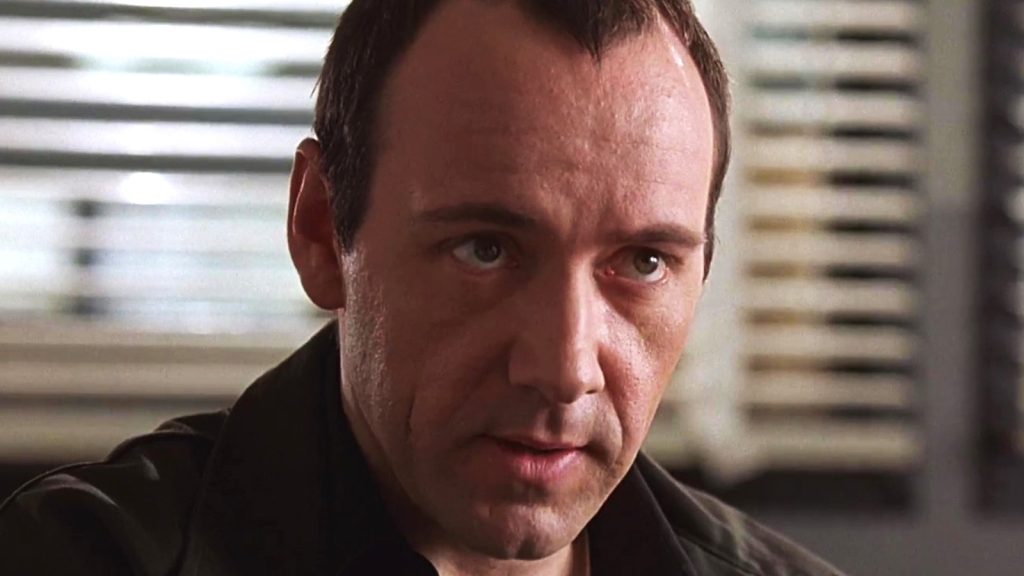 Close-up of a man with short dark hair and a wary, calculating expression, sitting in a dimly lit office with venetian blinds visible in the background. A still from The Usual Suspects (1995).