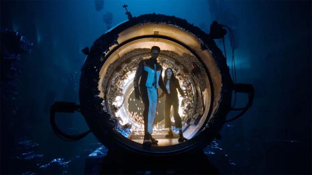 A man in a superhero suit and a woman stand together inside a glowing, circular pocket dimension portal, framed by a large metallic ring against a dark blue backdrop. A still from Superman (2025).