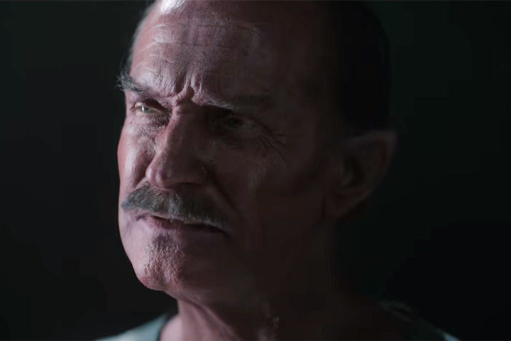 Close-up profile shot of a red-skinned, mustached older man with pointed ears and a stern expression, lit dramatically against a black background. A still from Lanterns (2026).