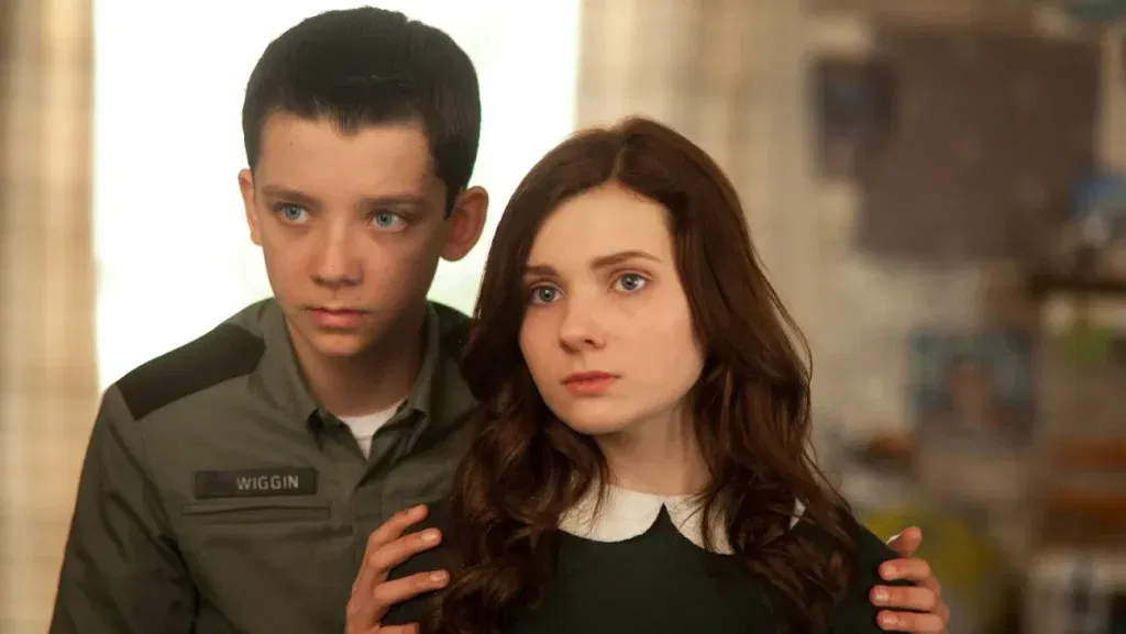 Asa Butterfield as Ender Wiggin, wearing a Battle School uniform with a "Wiggin" name patch, standing behind Abigail Breslin as Valentine Wiggin in a green sweater, her hand resting protectively over his.