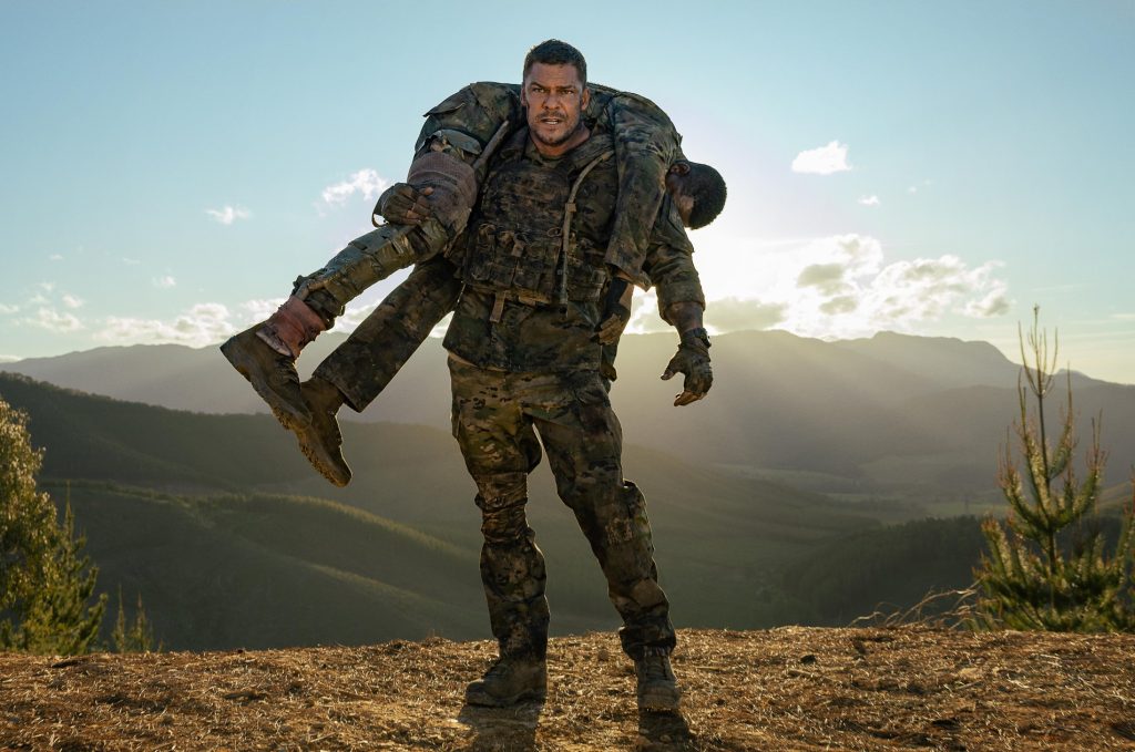 Alan Ritchson in muddy camouflage fatigues carries a wounded fellow soldier over his shoulder across a hilltop at sunrise, mountains visible in the background.