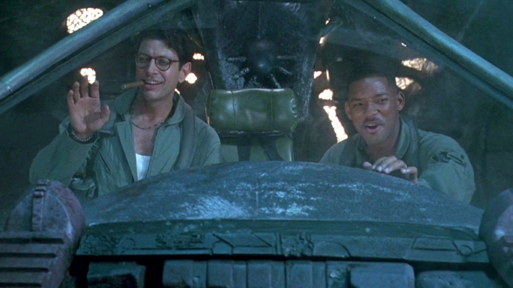 Jeff Goldblum smoking a cigar and waving beside Will Smith, both in flight jackets inside the cockpit of a captured alien ship, with the ship's occupant visible in shadow behind them.