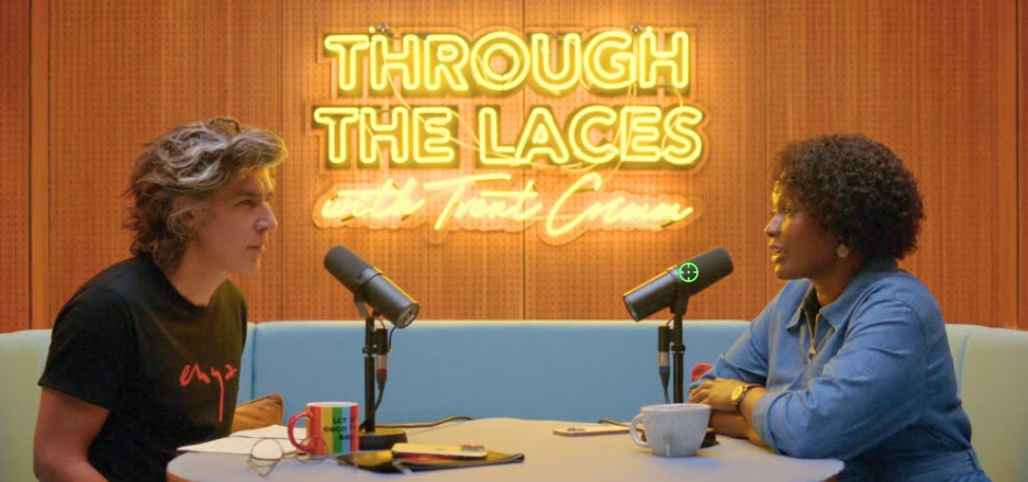 Two people sit across from each other at a round table with microphones in a warmly lit studio. On the left is a person with wavy gray-streaked hair wearing a black t-shirt; on the right is a person with short curly dark hair in a blue denim shirt. Behind them is a large yellow neon sign with Trent Crimm. A rainbow-striped mug and a plain white mug sit on the table.