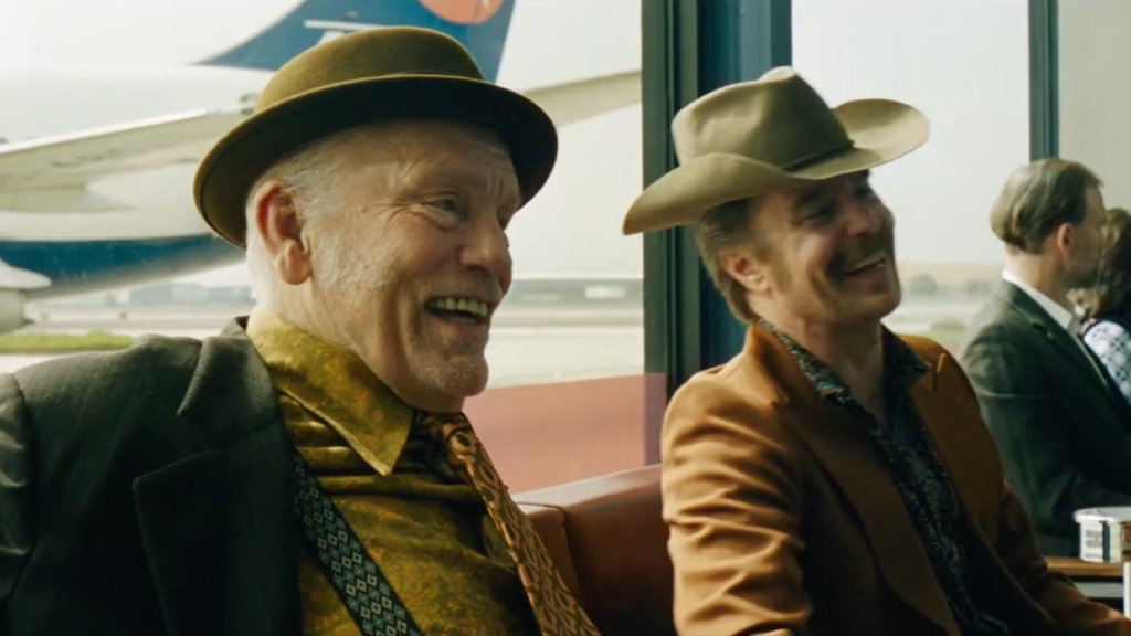 wo men wearing hats and retro clothing share a laugh while sitting in an airport terminal. An airplane is visible through the window behind them.