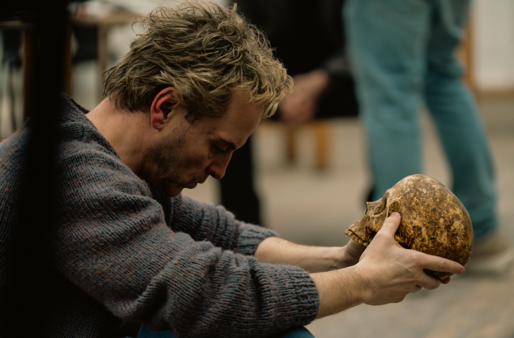A man in a thick knit sweater sits in profile, gazing intently at a weathered human skull he holds in both hands. The background is blurred, showing out-of-focus figures standing behind him.