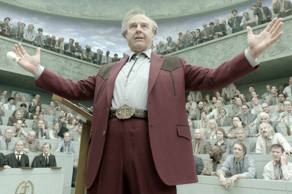 Tom Cruise, wearing heavy aging prosthetics, stands with his arms outstretched in a large, tiered amphitheater. He wears a maroon western suit, a bolo tie, and a large belt buckle, while an audience watches from the curved seating behind him.