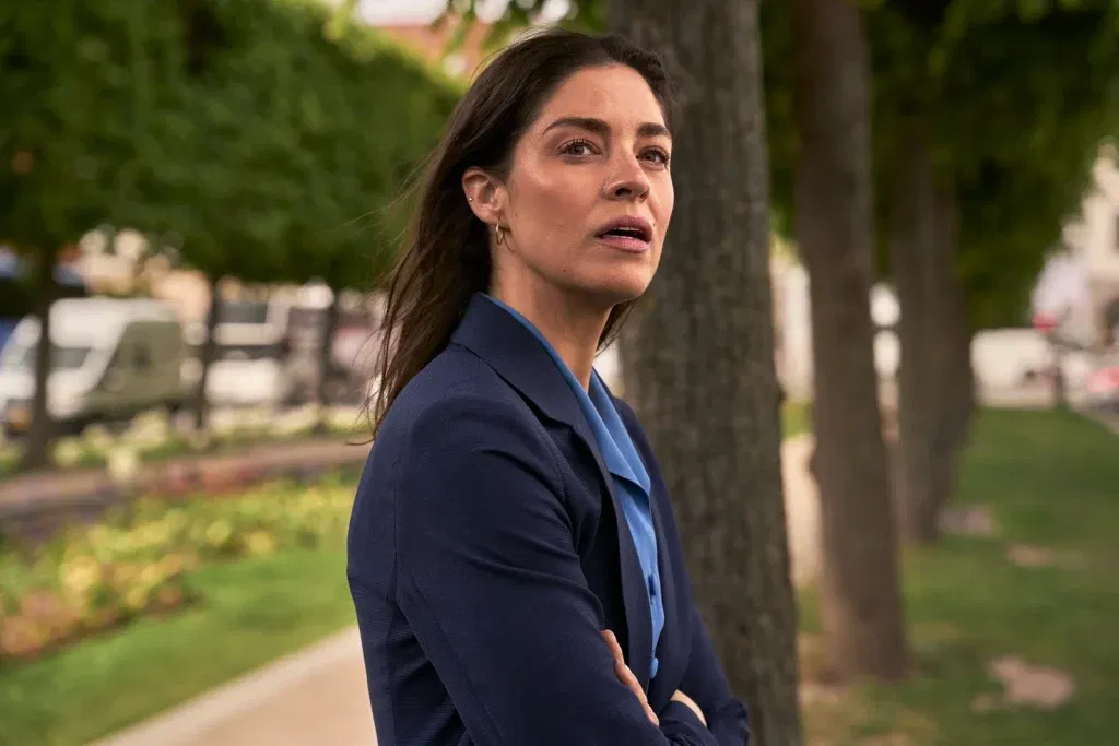 Natalie Madueño, as Louise/Helene in The Secret Woman, stands with arms crossed on a tree-lined city path, a startled, wary expression on her face.