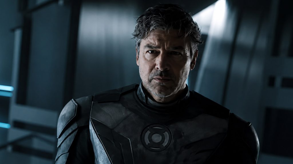 A grim, weathered man with graying, tousled hair stands in a dimly lit sci-fi corridor, wearing a dark, armored Green Lantern uniform with a glowing power battery symbol on the chest, illuminated by cool blue light from the sides.
