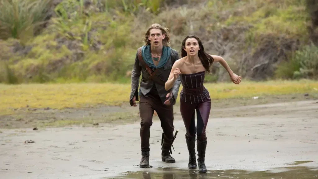 A young man with shoulder-length dirty-blond hair and a teal scarf runs alongside a dark-haired woman in a fitted burgundy corset top and leather leggings, both looking back over their shoulders with alarmed expressions as they flee across a muddy, overgrown landscape. The series is set on post-apocalyptic earth