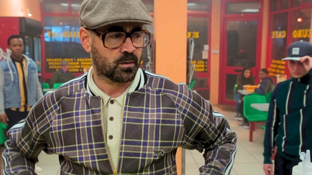 Colin Farrell as Coach in The Gentlemen, wearing a flat cap, thick-framed glasses, and a plaid track jacket over a cream polo, standing with hands on hips in a fast-food restaurant.