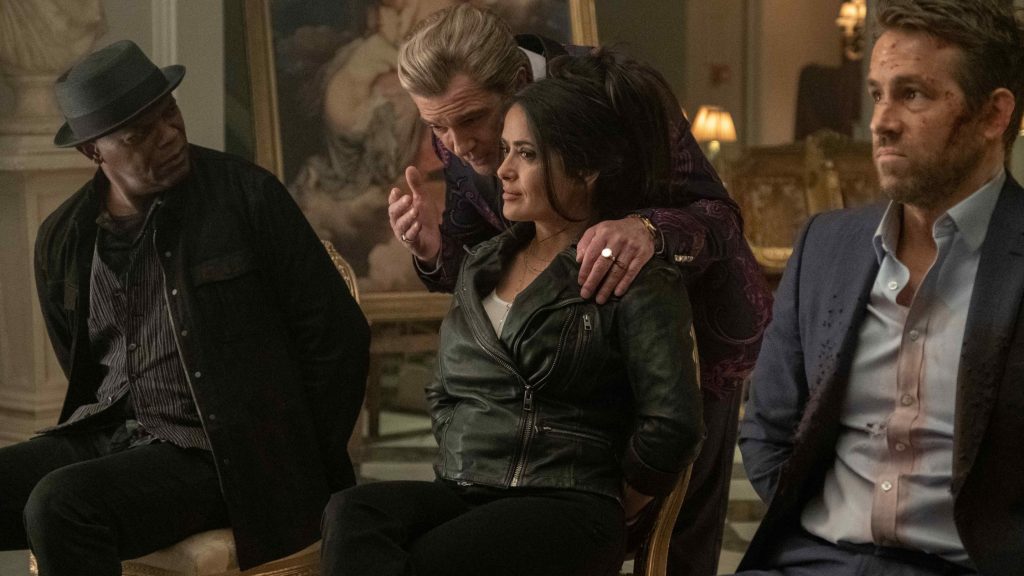 Four people seated in an ornate room. A man in a fedora watches warily on the left; center, a woman in a green leather jacket sits tensely as an older man in a paisley jacket leans over her shoulder; right, a bruised man in a rumpled blazer looks on tensely.