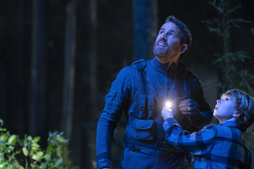 Ryan Reynolds in a tactical flight suit stands in a dark forest at night, lit in cool blue tones, gazing upward with a tense expression. Beside him, a boy in a plaid jacket holds a small glowing device and looks up in the same direction.