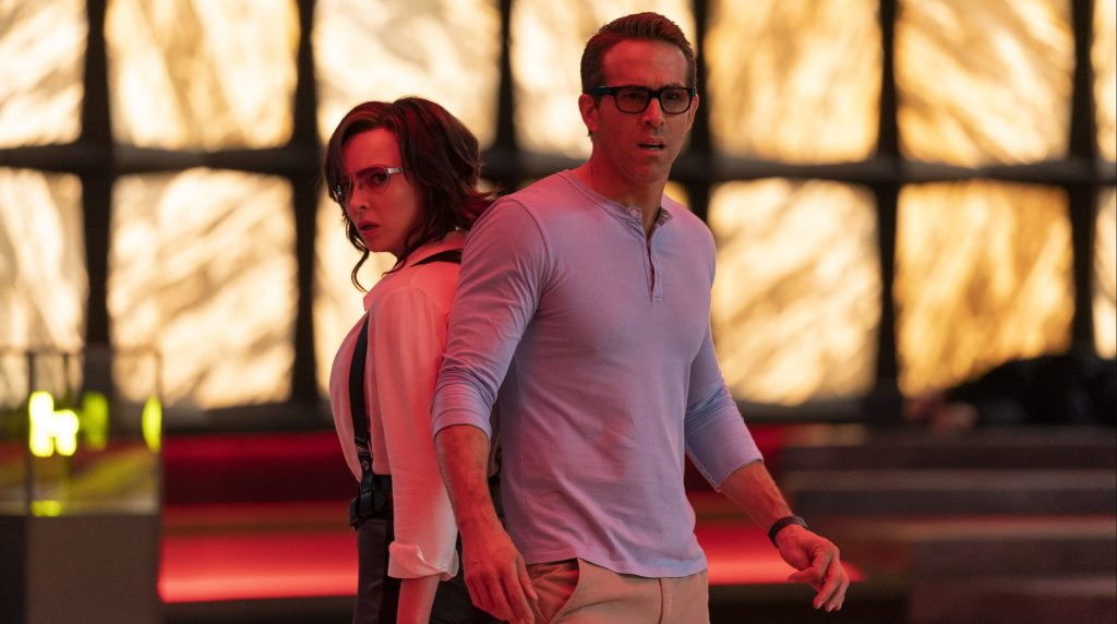 A man in glasses and a lavender henley stands in front, looking alert, while a woman with a shoulder holster over a white blouse looks back over her shoulder behind him, both lit in warm red-orange light against a glowing backlit wall.