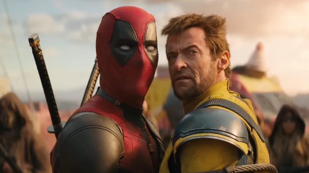 A close-up of two costumed superheroes standing side by side against a dusty, sunlit outdoor backdrop. On the left, a man in a red-and-black masked suit with sword hilts visible over his shoulder. On the right, a man with tousled hair in a yellow-and-blue armored suit, both looking off to the side with tense expressions.