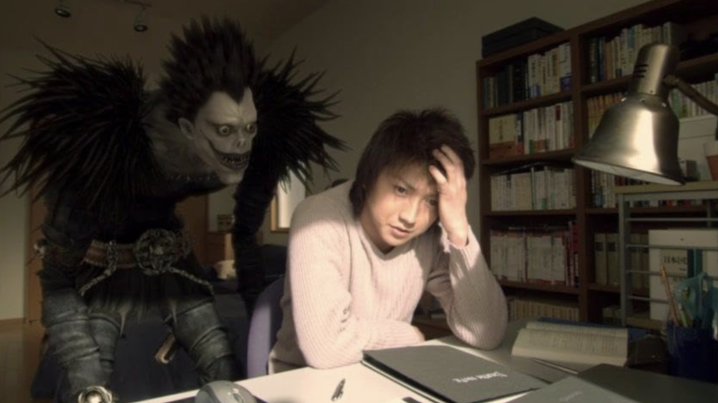 This image shows a scene from the 2006 live-action Japanese film adaptation of Death Note, featuring the character Ryuk standing behind a stressed student at a desk. 