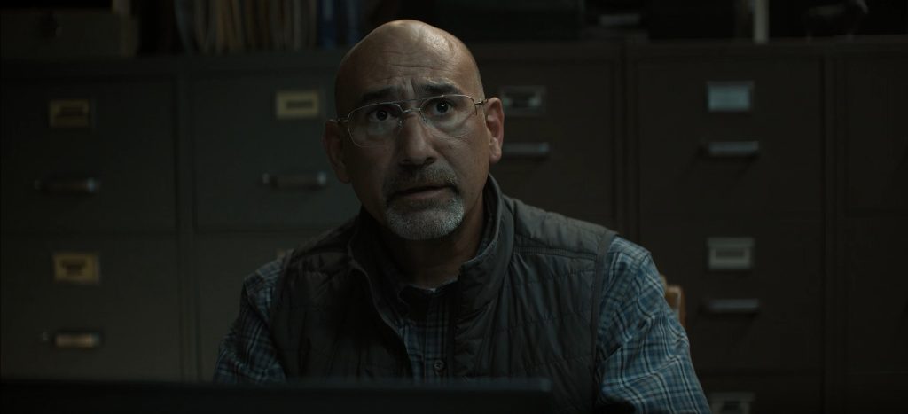 A bald, bespectacled man with a gray beard sits at a desk in a dimly lit office lined with filing cabinets, looking startled in Lanterns episode 4.