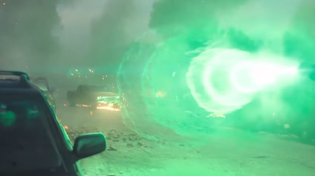 A bright green energy blast engulfs a car on a smoke-filled street, glowing brilliantly against the wreckage and sparks in the background.