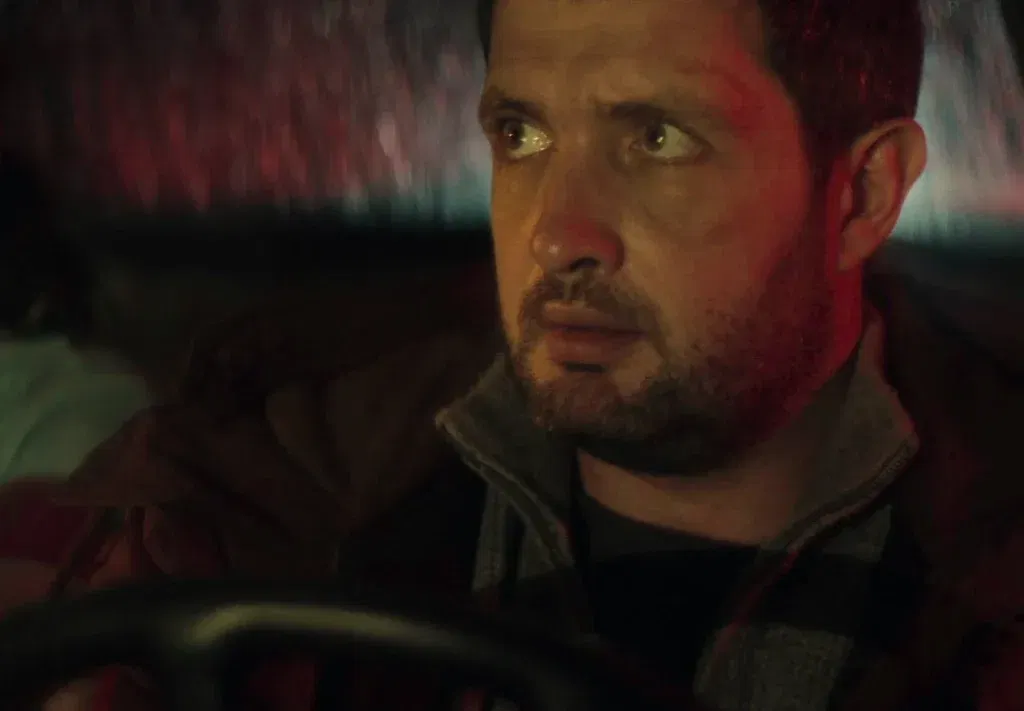 A bloodied man with a fresh cut above his eyebrow sits behind the wheel of a car at night, glancing anxiously to the side. Red and blue light washes across his face, and his expression is tense and wary.