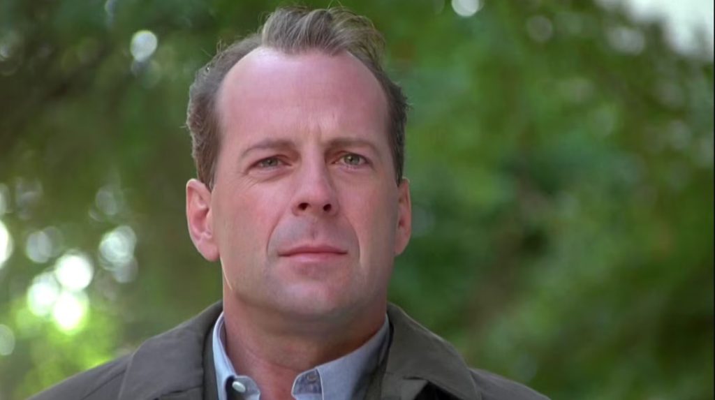 A close-up of a man in a light coat standing outdoors, his expression calm but distant, with a soft, out-of-focus green backdrop of trees behind him.