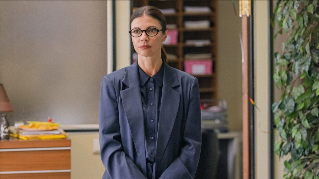 A woman with dark hair pulled back, round glasses, and a navy blazer over a matching shirt stands with a stern, appraising expression in an office hallway, with a mail sorting shelf, a hanging plant, and a plate of food on a cabinet visible around her, from Netflix's Go Team!.