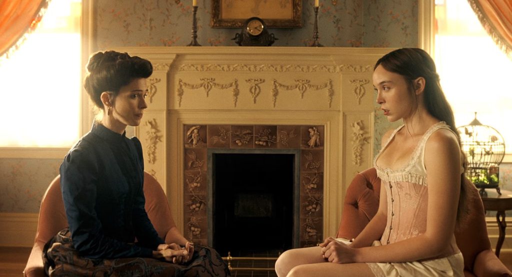 Two women sit facing each other on chairs beside an ornate fireplace, one in a dark blue high-necked Victorian dress, the other in a white lace corset and slip, in a sunlit parlor with a birdcage nearby.