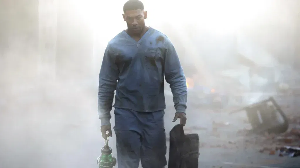 A man in dirt-stained blue scrubs walks through a smoky, debris-strewn area carrying a green lantern-like device and a dark duffel bag, harsh light glaring behind him.