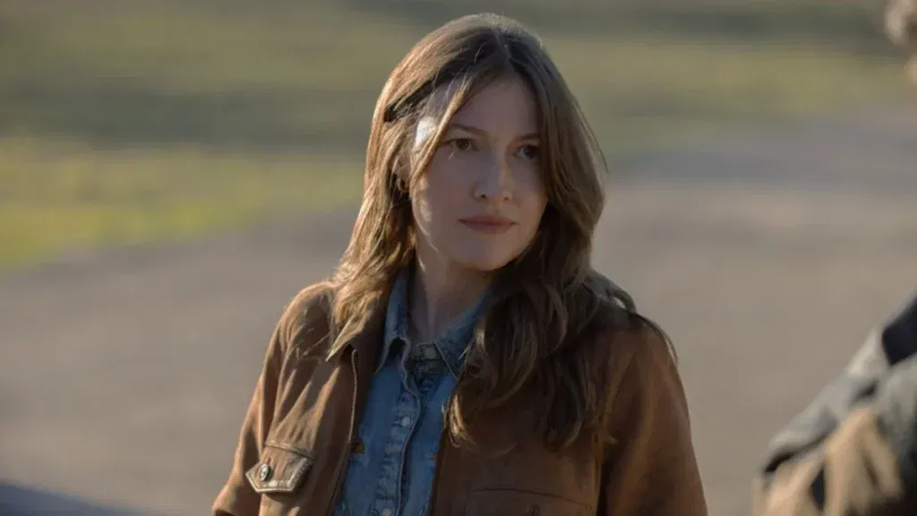 A woman with long wavy brown hair stands outdoors wearing a brown leather jacket over a denim shirt, her expression serious and watchful as she looks slightly off-camera, with a blurred rural landscape in the background. in lanterns episode 5 we learn more about her gotham origins.
