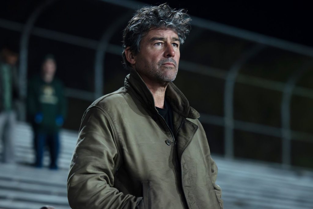 A man with tousled grey and dark hair and a stubbled face stands outdoors at night wearing an olive-green jacket, gazing off to the side with a serious, contemplative expression, with blurred figures and bleachers visible behind him.