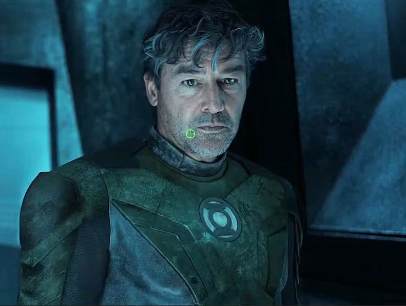 A rugged, weathered man with grey-streaked hair and stubble stands in a dim, industrial setting, wearing a worn green and grey suit with a glowing circular emblem on the chest.