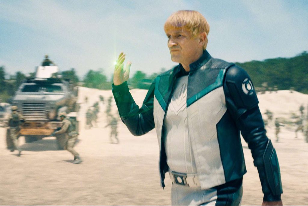 A blond man in a white and dark teal costume with a Green Lantern-style emblem on the shoulder stands in a sandy, sunlit outdoor setting, his hand raised with a small glow of green energy forming between his fingers, while soldiers and a military vehicle are visible blurred in the background.