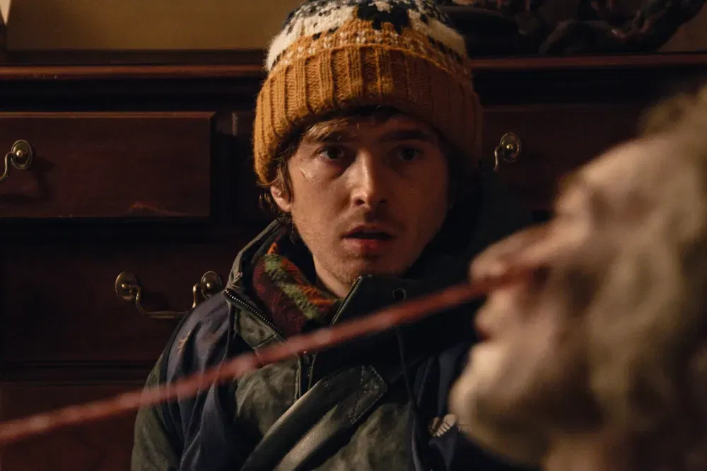 Resident Evil Ending Explained: What Does Bryan Become After Losing the Cure? 36 A close-up of a young man in a knit beanie and jacket, looking startled with a scratch on his cheek, as an out-of-focus pale, disheveled figure looms in the foreground.