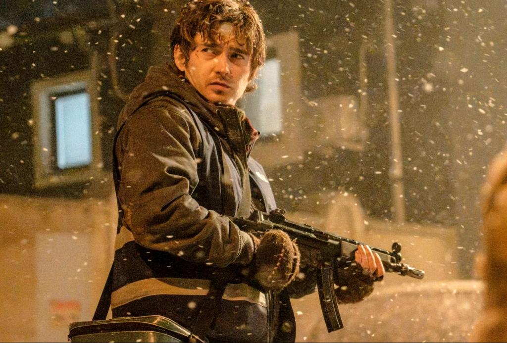 Resident Evil Ending Explained: What Does Bryan Become After Losing the Cure? 35 A young man with tousled wet hair braces a submachine gun while snow falls heavily around him at night, wearing a hooded jacket with a strap across his chest, standing in front of dimly lit buildings. A still from Resident Evil