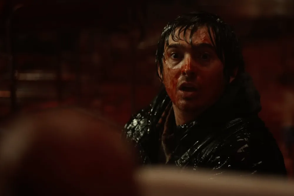 A wet-haired man with blood spattered across his face stares with a shocked, open-mouthed expression in a dark, red-lit interior, wearing a soaked black jacket, with an out-of-focus figure in the foreground.